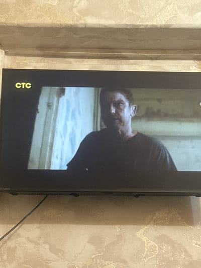 The image shows a television screen displaying a scene from a movie or television show. The logo "CTC" is visible in the upper left corner of the screen. The content on the screen features a man's face, partially in shadow, with a dark t-shirt. The background appears to be an interior with a window or opening with light coming through. The television itself is mounted on a textured, light-colored wall, likely wallpaper. A black cable runs from the bottom of the television down the wall. The bottom left corner of the television shows a logo that appears to be "SDER". The image is taken from a medium distance, capturing the television and the wall it is mounted on. There is no discernible indication of the city of Saransk, Russia within the image itself.