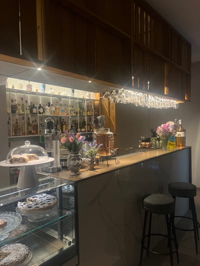 The image shows a dimly lit, modern bar area, likely a cafe or restaurant, in Vallo della Lucania, Italy. A display case in the foreground showcases several desserts, including a fruit tart and what appears to be a rolled pastry covered with powdered sugar. Behind the display case, a bar counter stretches across the frame. On the counter, a large glass dispenser filled with a pinkish beverage sits next to a vase of pink tulips and sprigs of lavender. Further back, shelves are stocked with numerous bottles of liquor and other beverages. Above the shelves, rows of wine glasses are suspended from the ceiling. Two dark upholstered bar stools with black legs are positioned in front of the counter. The overall ambiance is sophisticated and inviting, with warm lighting highlighting the products and decor.