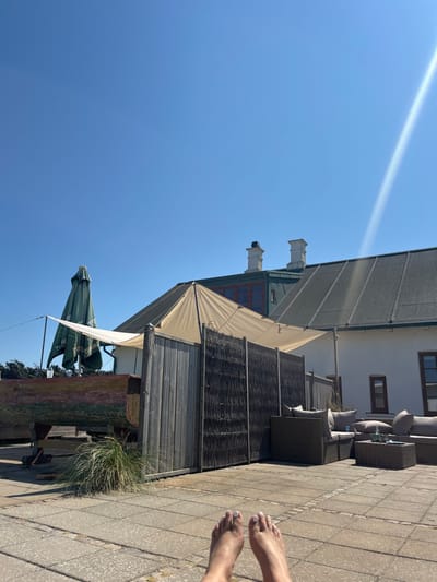 A low-angle shot captures an outdoor scene on a clear, sunny day in Skagen, Denmark. In the immediate foreground, a pair of bare human feet with white-painted toenails rests on light grey square patio pavers. The midground features an outdoor lounge area with dark brown wicker-style modular seating and light grey cushions on the right. A multi-panel privacy screen, composed of weathered vertical wooden planks and dark woven material, extends across the center. Above this screen, a beige fabric shade sail is tautly stretched. To the left of the screen, a large, weathered wooden boat hull, exhibiting traces of red and green paint, rests on wooden supports. A tall, folded green market umbrella stands behind the boat, partially obscuring "TUBORG" text. In the background, a white building with a dark grey sloped roof and multiple windows is visible. Two white chimneys rise from the roofline. The upper portion of the image is dominated by a bright, clear blue sky, with a pronounced sun flare extending diagonally from the upper right.