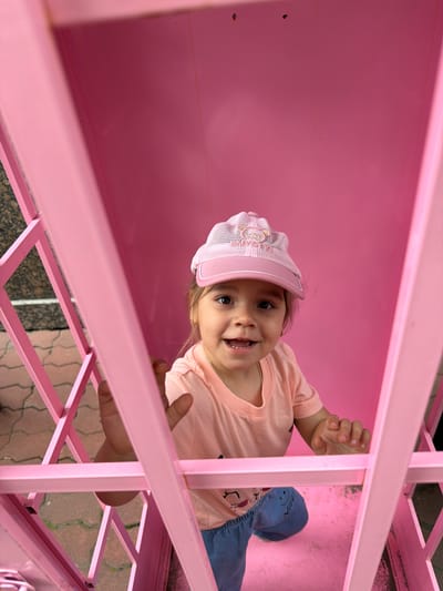 An overhead shot captures a young child wearing a pink mesh baseball cap and a peach t-shirt, smiling with their mouth open, showing two teeth. The child is positioned inside a pink, cage-like structure. The background consists of solid pink walls, and the floor appears to be a matching pink surface. The child's hands are visible, resting on the pink bars of the structure. The exterior of the structure shows a glimpse of a cobblestone sidewalk with pink and grey tiles.