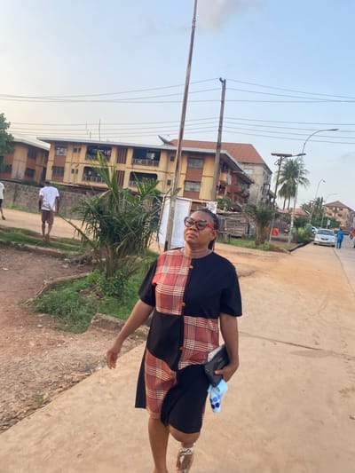 A woman is walking along a street in Abofifia, Nigeria. She is wearing glasses and a black dress with a red and brown plaid panel. She is carrying a black wallet, a white object, and a blue and white polka-dot object in her left hand. The street is paved and lined with buildings and trees. There is a person wearing a white shirt walking in the background. Power lines are visible overhead. The sky is light blue.