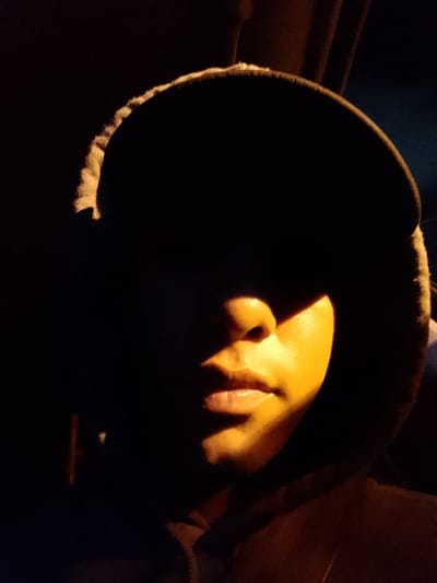 A photograph depicts a person in a hooded sweatshirt and baseball cap. The individual's face is partially illuminated by an orange light source, highlighting the nose and mouth. The rest of the face and the hood are shrouded in darkness. The setting is indeterminate. There is no information in the image to support a location in San Antonio, Venezuela.