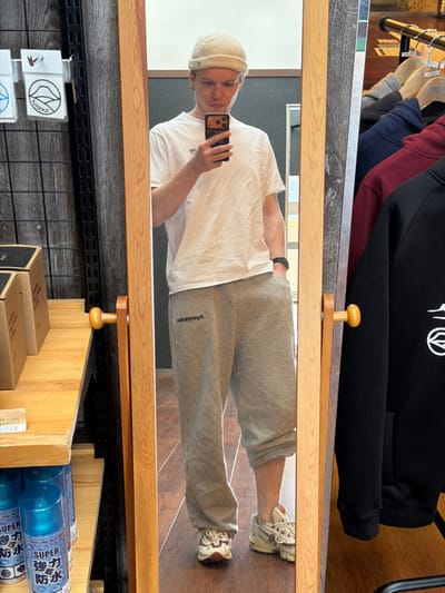 A mirror reflection shows a person wearing a white t-shirt, grey sweatpants, a light-colored beanie, and sneakers. The person holds a cell phone, presumably taking the photo. Above the mirror, hoodies are hanging on a rack. Below, shelves hold boxes and blue canisters with Japanese text. The mirror's frame is light wood with decorative knobs. The background includes a patterned wall and wooden flooring.