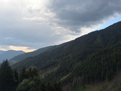 This media file depicts a scenic mountain landscape, likely taken during the late afternoon or early evening. The scene is dominated by rolling, forested mountains under a dramatic sky filled with dark, heavy clouds that are beginning to break, allowing patches of lighter sky and a hint of sunlight to filter through. The overall mood is serene and slightly somber due to the overcast conditions.
The foreground features the dark green silhouettes of evergreen trees, including several prominent fir trees. These trees are dense and appear lush. As the view recedes into the distance, the mountainsides are covered in a thick carpet of dark green pine trees, creating a rich texture across the slopes. There are visible signs of deforestation or logging in certain areas, with cleared patches and scattered dead, grey trees standing starkly against the live forest. The terrain is steep and uneven, characteristic of alpine environments.
In the mid-ground, layers of mountains recede into the distance, with the furthest peaks appearing as soft, bluish silhouettes against the hazy sky. A winding road is faintly visible on one of the slopes in the distance, suggesting human presence and accessibility to the area. The sky is a significant element, with a mix of dark, brooding storm clouds and lighter, brighter areas that suggest the sun is setting or has recently set beyond the horizon. The light is diffused, casting a soft glow on the landscape. No people, objects, or text are visible in the image. The location context provided is Freienfeld - Campo di Trens, Italy, which aligns with the mountainous, forested terrain.