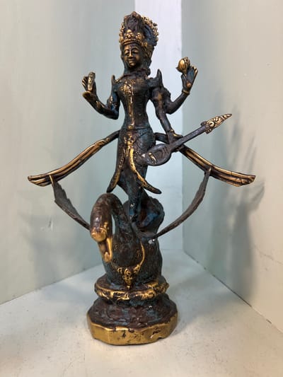 This is a photograph of a bronze statue. The statue depicts a figure with multiple arms, possibly a deity or mythical being, in a dynamic pose. The figure has intricate detailing on its clothing and jewelry, with golden accents highlighting its form. The statue is mounted on a dark, textured base that tapers upwards. It appears to be made of bronze with a patina that gives it an aged, antique look, with some areas showing hints of blue-green oxidation. The statue is positioned against a light-colored, textured wall and rests on a flat surface, casting a subtle shadow. The lighting is even, suggesting an indoor setting, possibly a display or storage area. The focus is on the detailed craftsmanship of the statue, which has a sense of movement and power. There is no visible text in the image.