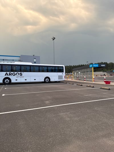 A white bus with "ARGOS LET'S GO!" written on its side is parked in a parking lot. The bus is positioned on the left side of the image, with its front end facing left. Behind the bus, a modern building complex with glass windows and a blue and white facade is visible. To the right of the bus is a bus stop shelter with a yellow frame and a blue sign that reads "Descuno" in Polish. The shelter is a covered waiting area with benches. The parking lot itself is paved with asphalt and marked with white lines. In the background, a dense forest of pine trees stretches across the horizon under a cloudy, overcast sky. The scene appears to be an outdoor area associated with a transportation hub or a large facility, possibly near Chociule, Poland, given the Polish signage at the bus stop. There are no people clearly visible in the immediate vicinity of the bus or the shelter.