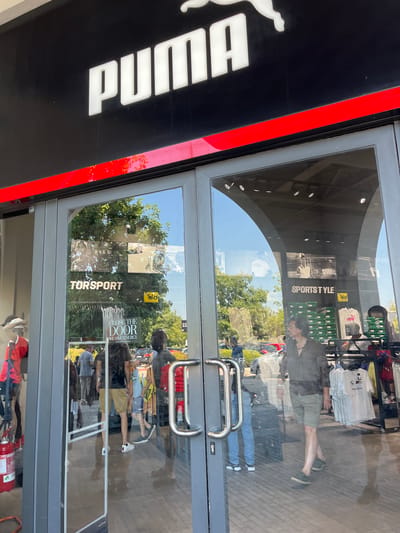 The image shows the exterior of a Puma store in Rodengo, Italy. The store has a black facade with the white Puma logo and name prominently displayed above the entrance. The entrance consists of large glass doors with silver handles. Reflected in the glass doors are trees, other buildings, and pedestrians. Inside the store, racks of clothing and shoe boxes are visible. A fire extinguisher is mounted to the left of the entrance. The overall scene depicts a typical retail environment on a sunny day.