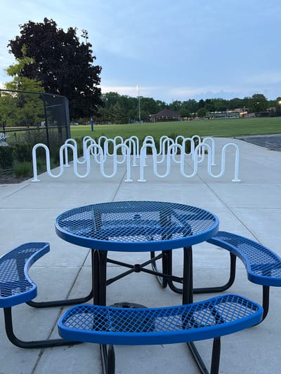 The image depicts a scene in Morton Grove, United States of America. A blue metal picnic table with attached benches sits on a concrete surface. Adjacent to the table is a row of white, serpentine-shaped metal bike racks. In the background, there's a grassy field, trees, and a fence. A black fence and dark leafy tree are in the foreground towards the left of the image. The sky is visible above the trees, suggesting dusk or early morning lighting.