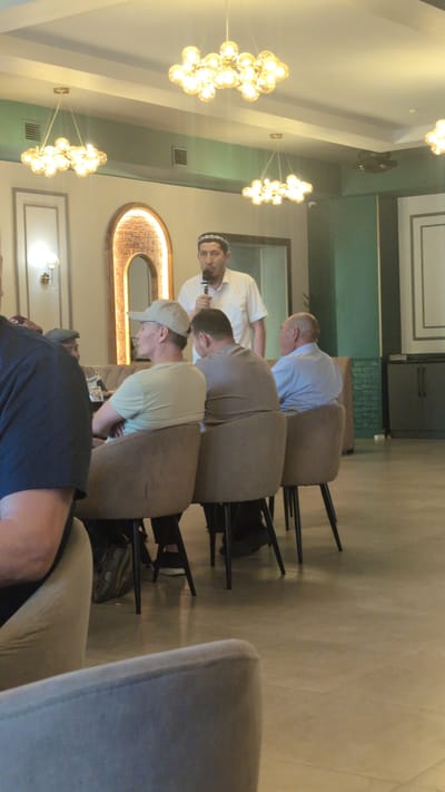 The image depicts a gathering of men indoors, possibly at a restaurant or event hall. A man in a white shirt and a traditional kufi cap is speaking into a microphone, standing while the others are seated. Several men are visible from the back, seated in chairs, facing away from the camera. The environment features modern lighting fixtures with multiple bulbs hanging from the ceiling, and the walls are decorated with subtle textures and framed elements. The floor appears to be tiled. The overall impression is of a formal or semi-formal assembly. The location is identified as Ötegen batyr, Kazakhstan.