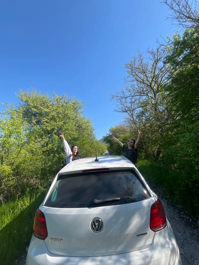 Two people are sticking their arms out of a white Volkswagen Polo car. The car is positioned in front of lush green trees with a clear blue sky above. The photo appears to be taken in a rural area near Rish, Bulgaria. The people are facing towards the car with their arms outstretched towards the trees. One person is wearing a grey sweatshirt and the other is wearing a dark colored shirt. The car is parked on a dirt road near grass.