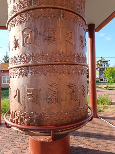 A large, cylindrical prayer wheel, made of bronze or copper, stands outdoors in the city of Verkhnyaya Ivolga, Russia. The wheel is elaborately decorated with intricate patterns and embossed with what appear to be Tibetan or Sanskrit script. It is supported by a reddish-brown base and a metal railing encircles its lower portion. In the background, a multi-story building with a distinctive pagoda-style roof is visible, along with trees and a bright blue sky. The ground around the prayer wheel is paved with red brick. The overall scene suggests a religious or cultural site, likely a Buddhist temple or monastery, given the presence of the prayer wheel and the architectural style of the building.