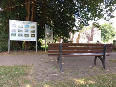 An outdoor informational sign displays the title "GEMEINDE ILLINGEN" and multiple small images of local points of interest, including historical buildings and landscapes. A wooden bench with dark metal legs is positioned in front of the sign, facing a stone ruin in the background, likely a historical structure within the municipality. Sunlight filters through the leaves of a large tree casting shadows on the paved walkway and surrounding grass. The overall scene suggests a park or historical site in Illingen, Germany, designed for visitors to learn about the area's attractions.