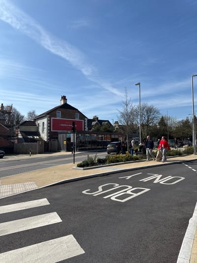 The image depicts a sunny street scene at the junction of Queen Street and Endless Street in Salisbury, United Kingdom. The sky is clear blue with faint, wispy cirrus clouds.
In the immediate foreground, a newly paved asphalt road features a white zebra crossing and the inverted white road marking "BUS ONLY". Adjacent to this, a light-colored block paving forms pedestrian pathways, bordered by a curb and raised planters containing small, sparsely leaved shrubs.
The central building is a two-story brick structure, identified as "The Queen's Head" pub, featuring a large red banner sign on its upper facade that reads "QUEEN'S HEAD BEERS & BURGERS". To the left of the pub, scaffolding is erected against an adjacent building. Beyond the main road, additional buildings and bare deciduous trees are visible. Modern, dark grey streetlights are positioned along the pavement on the right.
On the right-hand pavement, two pedestrians are walking away from the camera. The lead individual, a male, wears a dark blue long-sleeved top and light grey trousers, holding a white, circular container or tray in his right hand. Following him is a female, dressed in a red long-sleeved top and light grey trousers, carrying a light-colored bag or item in her right hand. Further in the mid-ground, several other individuals are standing near parked vehicles, which include a dark grey hatchback.