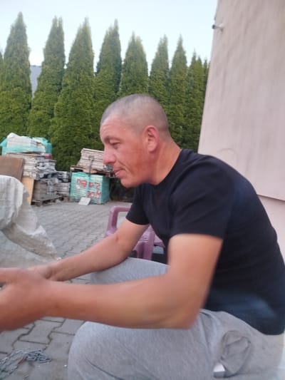 An adult male with a shaved head, wearing a black t-shirt and grey sweatpants, is seated outdoors. He is positioned in the foreground, facing towards the right side of the image and looking down and to his right. His arms are bent and resting in his lap, with his left hand appearing to hold something. In the background, several rows of tall, green evergreen trees are visible, lining a light-colored wall on the right. Piles of materials, including what appear to be stacked pallets and covered items, are situated in the middle ground. The ground is paved with grey interlocking bricks. A pink plastic chair is partially visible behind the man. The overall scene suggests an outdoor industrial or storage area. The image is assumed to be from Skórzewo, Poland.