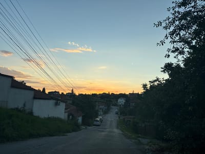 This image depicts a street scene in Ivanyane, Bulgaria, during sunset. The sky is a gradient of blue and orange, with a few scattered clouds tinged with yellow and pink. A cluster of residential buildings with red-tiled roofs lines the uphill street. Several cars are parked on the side of the road. Utility lines are visible stretching across the sky in the upper left portion of the frame. Lush green trees and vegetation flank the right side of the street and cover the hillside on the left. The overall impression is a tranquil, residential area bathed in the warm light of the setting sun.