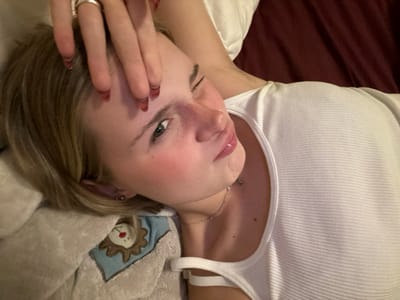 A young woman lies on her side, eyes squinted and lips pursed in a playful expression. She wears a white ribbed tank top and a delicate necklace. Her short, blonde hair is tousled. A hand, adorned with rings, rests on her forehead. She is nestled among plush pillows and blankets, one featuring a lion's head design. A dark, possibly maroon, surface is visible in the background.