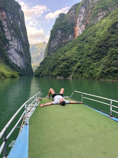 A person lies prone on the green AstroTurf deck of a boat, holding onto the metal railing with one hand. The boat appears to be traveling on a river or lake, with steep, forested mountains on the left side of the frame. The landscape suggests a location similar to the Đồng Văn Commune in Vietnam, characterized by rugged limestone karst formations covered in lush vegetation, and water. The boat's railing is made of white-painted metal, and the hull is painted light blue. The person is wearing a light-colored t-shirt, dark shorts, and sneakers. The water is dark green, and the sky is partly cloudy.