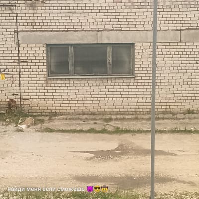 The image depicts an outdoor scene featuring a light-colored brick building with a row of three grimy windows. A thin, vertical gray pole is positioned in front of the building and extends upwards. The ground in front of the building appears to be a dirt or gravel surface with patches of grass and a puddle of water. Text overlaid on the bottom of the image reads "найди меня если сможешь" (find me if you can) in Cyrillic, followed by three emojis: a purple devil face, a yellow face with sunglasses, and a yellow face with a cowboy hat. The overall condition of the building and its surroundings suggests a utilitarian or industrial area. The specific location in Pskov, Russia, cannot be determined from the visual information alone.