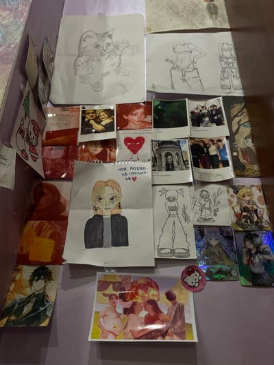 The image displays a wall covered with various items, including drawings, photographs, and what appear to be trading cards. The overall impression is one of personal display, possibly a bedroom or a creative space. The items are arranged in a collage-like fashion.
On the left side, there is a drawing of a cat. Below this are several photographs. One is a Polaroid-style photo of a couple embracing. Another shows a person's face in profile, appearing distressed. There are also images with text overlays.
In the center, a drawing of a person with blonde hair and a dark jacket is prominent. Above this drawing, there are more photographs, including one of a group of people outside a building and another of a couple. A drawing of a heart with a face is also visible.
To the right, there are drawings of figures in casual attire, and several trading cards featuring anime-style characters. A circular Hello Kitty sticker is also present.
The background wall is a solid, muted color, possibly purple or lavender. The lighting suggests an indoor environment, possibly with artificial light sources contributing to the overall ambiance. There is no visible indication of the specific location within the city of ЖК «Борисоглебское», Russia. The image focuses solely on the decorated wall.