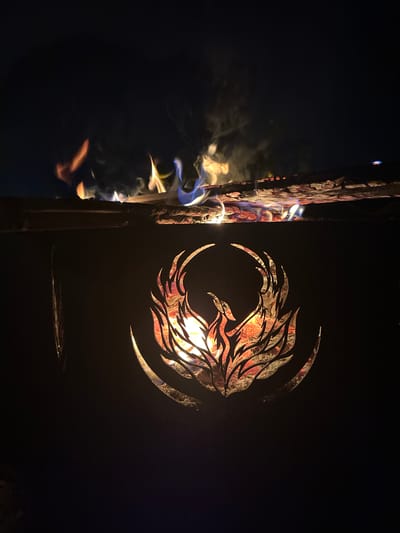 The image captures a nighttime scene featuring a burning fire, likely within an outdoor heating structure or fire pit, set in Веревское, Russia. The primary subject on the left is a dark, possibly metallic, panel with an intricate, decorative cutout. This cutout forms a swirling, abstract pattern resembling wings or stylized flames, through which bright, warm firelight emanates. The light visible through the cutout displays vibrant hues of orange, red, and yellow.
To the right of this panel, a thick, vertical wooden element, possibly a log or a structural part of the fire feature, is actively burning. Licks of fire, exhibiting colors from bright orange and red to cooler blues and whites, cling to and consume the wood. White, translucent smoke billows upwards from the burning section, adding to the dynamic nature of the flames.
The overall setting is dark, emphasizing the intense luminosity and colors of the fire against the black background, indicative of nighttime. No people are visible, and there is no discernible text. The image highlights the warmth and visual appeal of a contained outdoor fire.