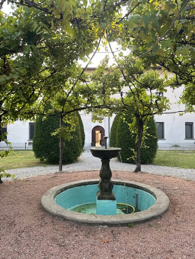 In Balerna, Switzerland, a small, circular fountain with a turquoise interior sits in the foreground, surrounded by reddish-brown gravel. Water flows from a dark stone pedestal into the fountain. Behind the fountain, two large, cone-shaped bushes flank a building with white walls and arched doorways. A vine-covered pergola arches over the scene, with green leaves and some clusters of grapes visible. The building has several windows with dark grates. The overall setting appears to be a tranquil courtyard.
