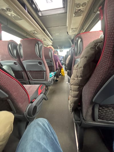 This image depicts the interior of a passenger coach bus, specifically in Dobrodan, Bulgaria. The perspective is from a seated passenger looking forward down the central aisle. The bus features rows of red and black fabric seats with gray plastic backs and integrated armrests. Overhead compartments with recessed lighting and air vents line the ceiling, which also includes a rectangular skylight.
In the foreground, the legs of the person taking the photo are visible, wearing light blue corduroy pants on the left. Adjacent to this, to the right, are the legs of another individual wearing beige pants. A brown puffer jacket hangs over the back of the seat directly in front and to the right of the camera's position. Further down the aisle on the left, a bottle of water with a blue cap rests in the mesh pocket of a seat back. Beyond this, a passenger wearing a purple top is visible, holding a yellow plastic bag in their hands. Other passengers are seated further toward the front of the bus, their figures partially obscured. The aisle floor is a uniform dark gray.