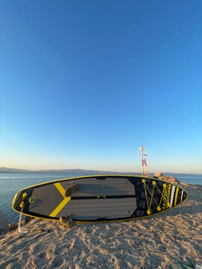 A stand-up paddleboard rests on a rocky shoreline with a bright blue sky overhead. The paddleboard is gray and yellow with black accents and bungee cords on the front. In the background, a navigation marker stands on the rocks, and a body of water stretches to the horizon, meeting distant hills. The scene appears to be set in Nea Potidea, Greece, likely during daylight hours.