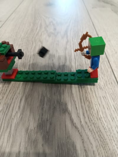 The image features a scene of two LEGO figures on a light-colored wooden floor. On the left is a structure made of brown and green LEGO bricks, resembling a small fort or barricade, with a black LEGO piece extending from its front. In the center, a small black LEGO cube is in mid-air, suggesting it has been launched or dropped. To the right, a LEGO minifigure with a green block head, blue torso, and red legs is holding a brown LEGO bow and arrow, appearing to be in the process of shooting or aiming. The minifigure has white hands. The scene is set indoors on a wooden surface that appears to be a floor with a visible grain pattern. The lighting suggests daytime, but there are no explicit cues about the weather or a specific time of day. There is no visible text in the image. The overall context suggests a playful or action-oriented scenario created with LEGO toys.