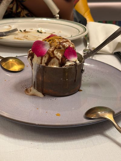 A tilted close-up shot shows a dessert presented in a cylindrical metal container, likely in a restaurant setting. The dessert appears to be a layered creation, possibly ice cream or mousse with chocolate sauce, topped with crushed nuts and adorned with two pink rose petals. The container sits on a light purple plate, which has a few crumbs scattered on it. Two spoons, one silver and one gold, are positioned on the plate. The setting includes other plates, a tablecloth, and hints of other diners and furniture in the background. The scene is indoors, well-lit, and suggestive of a formal or upscale dining experience. There is no specific information or visual clues that directly connect the image to the city of Qinhuai District, China.