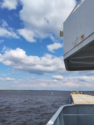 The image captures a view from a vessel on a wide body of water, likely a river, under a sky filled with scattered white clouds against a bright blue backdrop. In the foreground, a portion of a ship's deck and superstructure is visible on the right side, showing a weathered white surface with streaks of rust. The water is dark and choppy, suggesting a moderate breeze. In the distance, a thin strip of green land borders the horizon, indicating a coastline. A small white sailboat can be seen on the water in the mid-ground. Other vessels are faintly visible on the horizon. The scene is a static, outdoor environment.