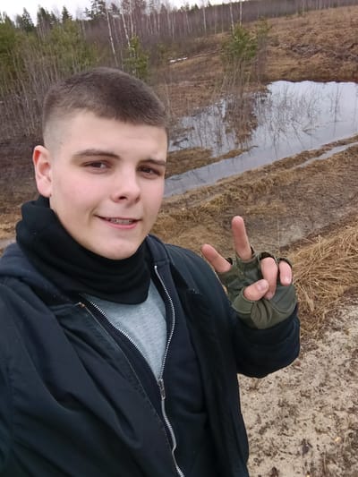 A selfie of a young man with short, light-brown hair is taken outdoors. He is wearing braces, a black jacket, a gray shirt, a black neck warmer, and green fingerless gloves. He is giving a peace sign with his gloved hand.
The background shows a body of water, likely a swamp or large puddle, reflecting trees and the overcast sky. The terrain is brown and muddy, with dry grasses visible. In the distance, there are bare trees and some evergreens.
Based on the information provided, this photo was taken in Krasnyi Mayak, Russia. The specific location within the city is not identifiable from the image.