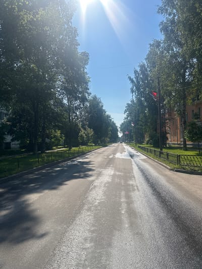 The image depicts a straight, paved road lined with mature trees on both sides. The road surface appears somewhat wet and reflective, suggesting recent rain or a street cleaning. On the right side of the road, there is a black metal fence bordering a grassy area and buildings. Several flags, red and white, are visible on poles along the street. The sky is bright blue with a strong sun, causing lens flare in the upper left corner. The scene is likely a residential or public street in Posyolok Imeni Morozova, Russia, captured on a sunny day. There are no people or vehicles clearly visible in the immediate foreground, though a vehicle can be seen in the distance down the road.