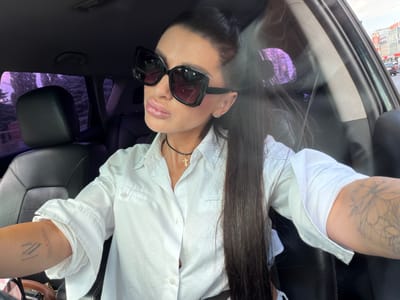 The media file captures a woman seated in the driver's seat of a car. She is wearing large, black-framed sunglasses and a white collared shirt, unbuttoned at the top, revealing a thin black choker with a golden cross pendant. Her long, dark hair is pulled back, with some strands falling forward over her shoulder. Tattoos are visible on her right arm. The background shows the interior of a car, with a black leather seat and a glimpse of the exterior through the window, which includes trees and buildings. The lighting suggests it is daytime, possibly late afternoon, as indicated by the bright sky and street scene visible outside. Reflections on the window suggest a somewhat cloudy or hazy sky. The overall mood appears casual and confident. The visible text includes a partial sign in the background, appearing to be Cyrillic script. The provided location context is Александровка, Russia.