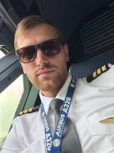A male pilot is depicted in a selfie taken inside the cockpit of an Airbus A320 aircraft, likely at London Gatwick Airport in Crawley, United Kingdom. The man, appearing to be in his late 20s or early 30s, has light brown hair and a neatly trimmed light brown beard. He is wearing dark brown Ray-Ban sunglasses. His uniform consists of a white pilot's shirt, a grey patterned tie, and black epaulets with four gold stripes, indicating the rank of Captain. A gold pilot's wing pin is affixed to his left chest pocket. Around his neck is a blue lanyard with the text "AIRBUS A320" and "AN EADS COMPANY" prominently displayed. The background shows the interior of the cockpit, with a window visible on the left side featuring light reflections and what appear to be raindrops. A dark control panel is partially visible on the right, and the pilot is seated in a dark blue seat with a striped pattern.