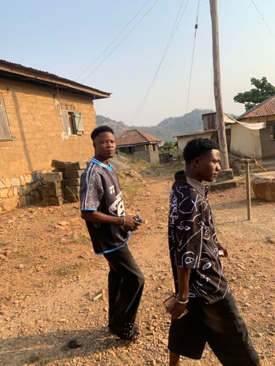 Two young men stand in a rural, sunny setting. One wears a black and white patterned shirt, black pants, and holds a phone. The other sports a black and gray jersey with black pants, also holding a phone. A modest brick building with closed windows stands to their left. In the background, a hillside is visible, along with a few simple structures. The ground is dry and covered in small rocks and gravel. A utility pole stands nearby.