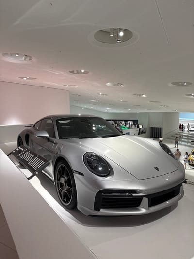The image shows a silver Porsche 911 Turbo S on display in the Porsche Museum in Stuttgart, Germany. The car is positioned on a white pedestal in a well-lit exhibition hall with a modern, minimalist design. The ceiling features a grid of recessed circular lights and ventilation grilles. A black informational plaque is placed in front of the car, providing details about its model and specifications. In the background, several other vehicles are visible, along with displays and architectural elements of the museum. A few museum visitors are also present, looking at the exhibits.