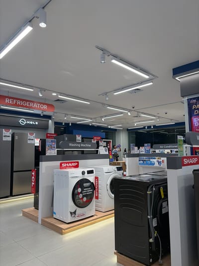 This is a horizontal, eye-level shot of an electronics store in Pekalongan, Indonesia. The store is well-lit with numerous fluorescent and spotlight fixtures. The scene is dominated by various home appliances, including refrigerators and washing machines, prominently displayed on shelves and stands. Brands like "SHARP" and "TCL" are clearly visible on product packaging and signage. A sign indicates "Air Conditioner" and another "Washing Machine." The overall atmosphere is that of a retail environment, with products neatly arranged for sale. The lighting suggests it's daytime, and the indoor setting implies controlled weather conditions. There are no people clearly visible in the image, but a person can be seen in the background, partially obscured. The floor is tiled, and the walls are painted white and dark blue. The text in the image includes brand names, product types, and promotional phrases like "POWER SAVING KING" and "Hello, Comfort."