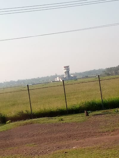 A distant view of an airport control tower amidst a vast grassy field, secured by a chain-link fence. The tower is a tall, multi-tiered structure with a prominent cabin on top. Adjacent to the tower are low-lying buildings. The foreground shows a dirt path with patches of grass. Overhead, power lines stretch across a hazy sky.