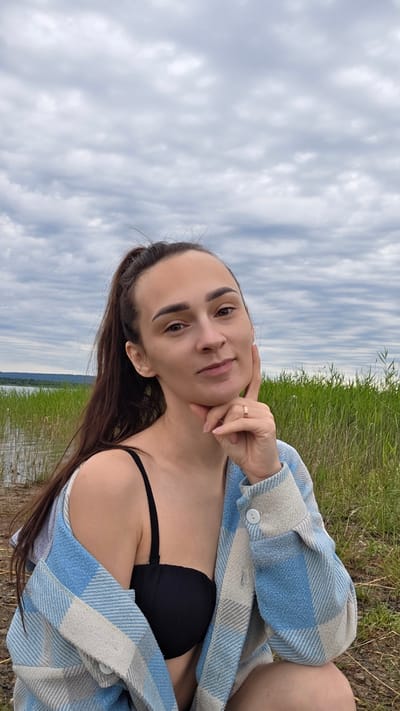 A young woman with dark brown hair pulled into a ponytail is captured in a medium shot, posing outdoors. She is wearing a black bikini top and a light blue and white checkered open shirt. Her right hand rests against her chin, and she wears a gold ring on her ring finger. The background consists of tall green reeds bordering a body of water, with a distant shoreline visible under a cloudy sky. The scene appears to be set in a natural environment, possibly near a lake or river.