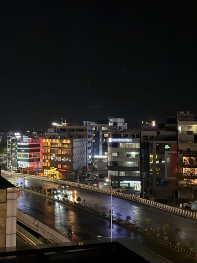 The image shows a night view of a wet, multi-level road in Shamshabad, India, with several buildings illuminated by artificial lights. The road, likely an elevated highway or flyover, has a dark asphalt surface reflecting the lights from the buildings and vehicles. Cars and a motorcycle are visible on the road, their headlights and taillights creating streaks of light due to motion blur and reflections.
Along the sides of the road are concrete barriers with black and white stripes, and planters with small trees and shrubs. The buildings are of varying heights and architectural styles. Some buildings have prominent signage, such as "TRIDENT HOSPITALS" and "ROYAL ENFIELD". The windows of the buildings are lit, indicating activity within. The sky is dark, suggesting it is nighttime, and a few distant lights can be seen in the background, possibly indicating the spread of the city. The overall scene depicts a busy urban environment at night, with the wet road adding a dynamic element to the cityscape.