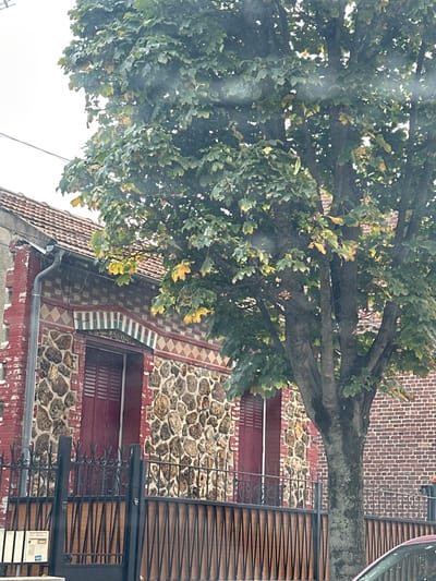 This image depicts a static street-level scene in Stains, France. The primary subjects are a large deciduous tree and a section of a building's facade, viewed under an overcast sky.
A prominent multi-trunked tree with dense green foliage, interspersed with yellowing leaves, occupies the right half of the frame. To its left, a portion of a building is visible. The building's lower facade is constructed from irregularly shaped, light-colored stones set in mortar. Above this, a decorative band features red brickwork with white diamond patterns, culminating in an arched section of vertical green and white tiles above two openings. These openings are fitted with closed, red louvered shutters. The building's roof, partially visible on the upper left, is composed of terracotta tiles, with a grey metal gutter and downspout running along its edge. The corner of the building on the far left shows exposed red brick.
In the foreground, a black wrought-iron fence with a solid dark panel beneath it extends across the scene, acting as a barrier. A white utility box or mailbox is affixed to the leftmost post of this fence. No individuals or observable activities are present. A red vehicle's bodywork is partially visible in the bottom right corner of the frame.