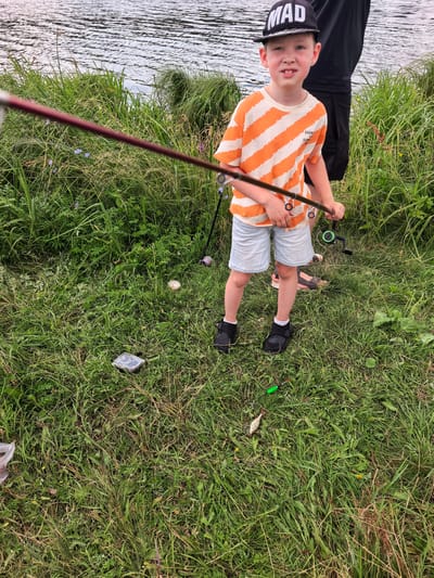 In the village of Kuzminovka, Russia, a young boy stands near a body of water, holding a fishing rod. He wears a striped orange and white t-shirt, light blue shorts, black shoes with white socks, and a black baseball cap with the word "MAD" in white letters. To his right, another person, partially visible, is also present. The ground is covered in lush green grass, with various plants and wildflowers growing. In the grass near the boy, a small fish and a green fishing float are visible. A dark rectangular object, possibly a container, is also on the ground. The water's surface has gentle ripples, reflecting the sky.