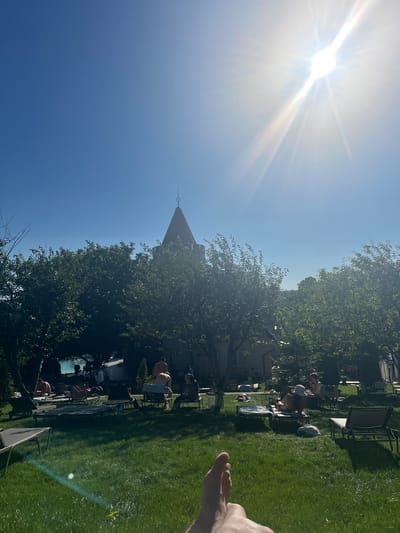 The image depicts an outdoor scene at a resort or hotel in Stăuceni, Moldova, on a sunny day. In the background, a distinctive turreted building with a red roof is visible, partially obscured by lush green trees. The foreground features a well-maintained lawn with several lounge chairs, many of which are occupied by people sunbathing or relaxing. Some individuals are visible near a swimming pool, suggesting recreational activities. The sky is clear and bright blue, with the sun prominently displayed, causing lens flare. The overall atmosphere is one of leisure and outdoor enjoyment.