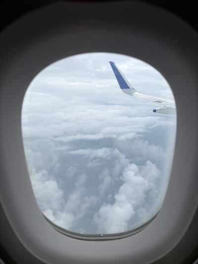 The image shows an airplane wing with a blue tip visible through an oval-shaped airplane window. Below the wing is a vast expanse of clouds. The exterior of the window frame is dark and out of focus, with the interior of the frame appearing as a light gray oval. The scene suggests an aerial view from an airplane. The provided location information, "Lakhiwali, India," is not visually verifiable from the image itself, as the image only depicts clouds and an airplane wing.