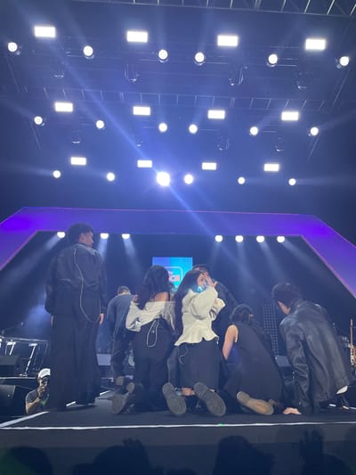 A group of performers bow on a stage with bright stage lights. The stage has a purple border. The performers wear dark clothing with some white accents. A camera operator is on the right side of the stage. The stage is in Salembaran, Indonesia.