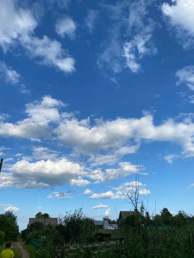 The image depicts a bright, sunny day with a clear blue sky filled with fluffy white clouds. The scene is oriented vertically, suggesting it was taken in portrait mode. On the right side of the image, a lush green landscape is visible, with dense trees and foliage. Scattered amongst the greenery are hints of human habitation, including what appears to be a modest building with a corrugated roof and possibly a greenhouse. A dirt path winds through the vegetation, leading the eye deeper into the scene.
In the foreground on the right, the back of a person wearing a bright yellow shirt is visible, their head and shoulders showing as they look away from the camera. The overall impression is one of a rural or semi-rural setting, possibly a backyard or a garden area in a village. The time of day appears to be daytime, given the brightness of the sky and the shadows cast. The weather is fair and pleasant, with the clouds adding visual interest to the sky. The location context provided places this scene in Novyi Ural, Russia, which aligns with the rustic and natural elements observed. There is no visible text in the image.