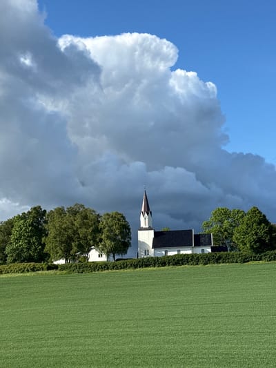 The image shows a white church with a dark roof and steeple, situated in a green field with trees in the background. The sky is partly cloudy with a bright blue sky visible in the upper right portion of the image. The church appears to be a traditional architectural style with arched windows and a pointed steeple. There are no people visible in the image. The scene is serene and picturesque, typical of a rural landscape in Norway. The location is identified as Kråkstad, Norway, suggesting a historical or religious site within that region. The overall atmosphere is tranquil and the natural setting is well-maintained, with a neatly trimmed hedge separating the church and trees from the foreground field.