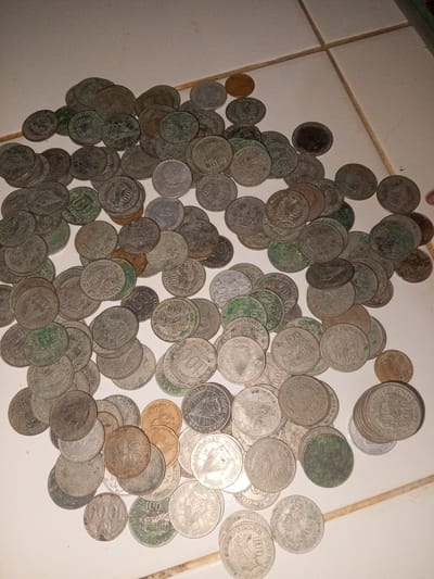 A close-up overhead shot shows a collection of Indonesian coins scattered on a white surface. The coins vary in color from tarnished silver to shades of copper and green. Many appear to be 100 Rupiah coins, with legible markings like "BANK INDONESIA," "SERATUS RUPIAH," and "100." Other visible denominations include 500 Rupiah and 1000 Rupiah coins. The coins are arranged somewhat randomly and overlap. The surface they rest on is slightly textured and has beige grout lines, indicating it is likely a tiled surface. There is no indication or information about the city of Kruenglingka Barat, Indonesia.