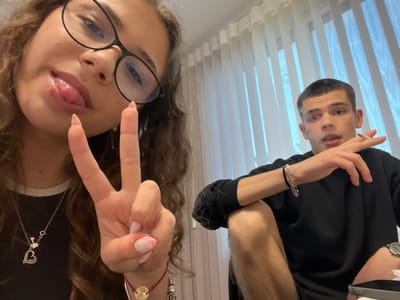 Two people are in a vertically oriented selfie image. One person is male, fair-skinned, with short dark hair, dark eyebrows, brown eyes, and is wearing a black top. He is positioned on the left, looking at the camera. The other person is female, fair-skinned, with brown curly hair, wearing glasses and a dark top with a white collar. She is making a peace sign with her right hand near her face and sticking out her tongue. In the background, there are vertical blinds. The location, Karavelovo, Bulgaria, is not discernable from the image.