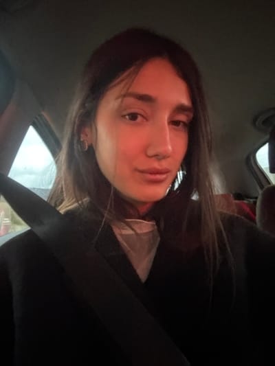 A person with long dark hair is sitting in a car, wearing a seatbelt. They are looking directly at the camera with a slight smile. The background is partially visible through the car windows and is out of focus. The interior of the car is dark, with the seatbelt prominently displayed across the frame. The lighting creates shadows on the person's face.
